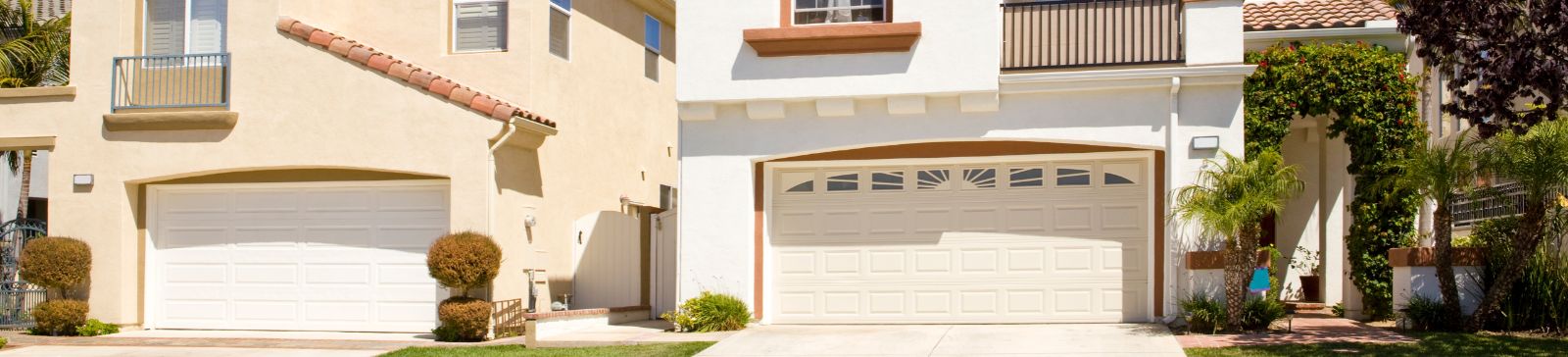 Two contemporary stucco houses with white sectional garage doors, red tile roofs, and manicured landscaping under a clear blue sky.